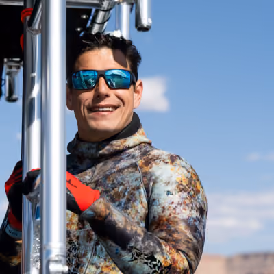 Smiling man in a camouflage wetsuit and sunglasses climbing metal ladder on a boat under blue sky.