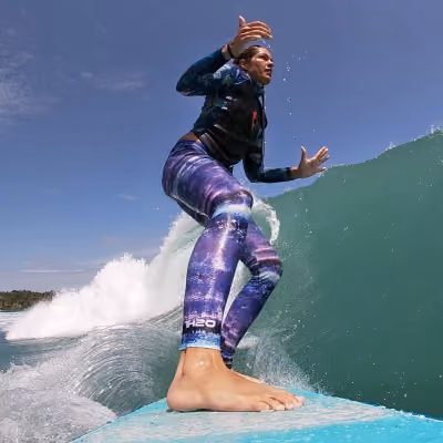 Person surfing a wave wearing galaxy-patterned leggings and a black wetsuit against a clear sky.