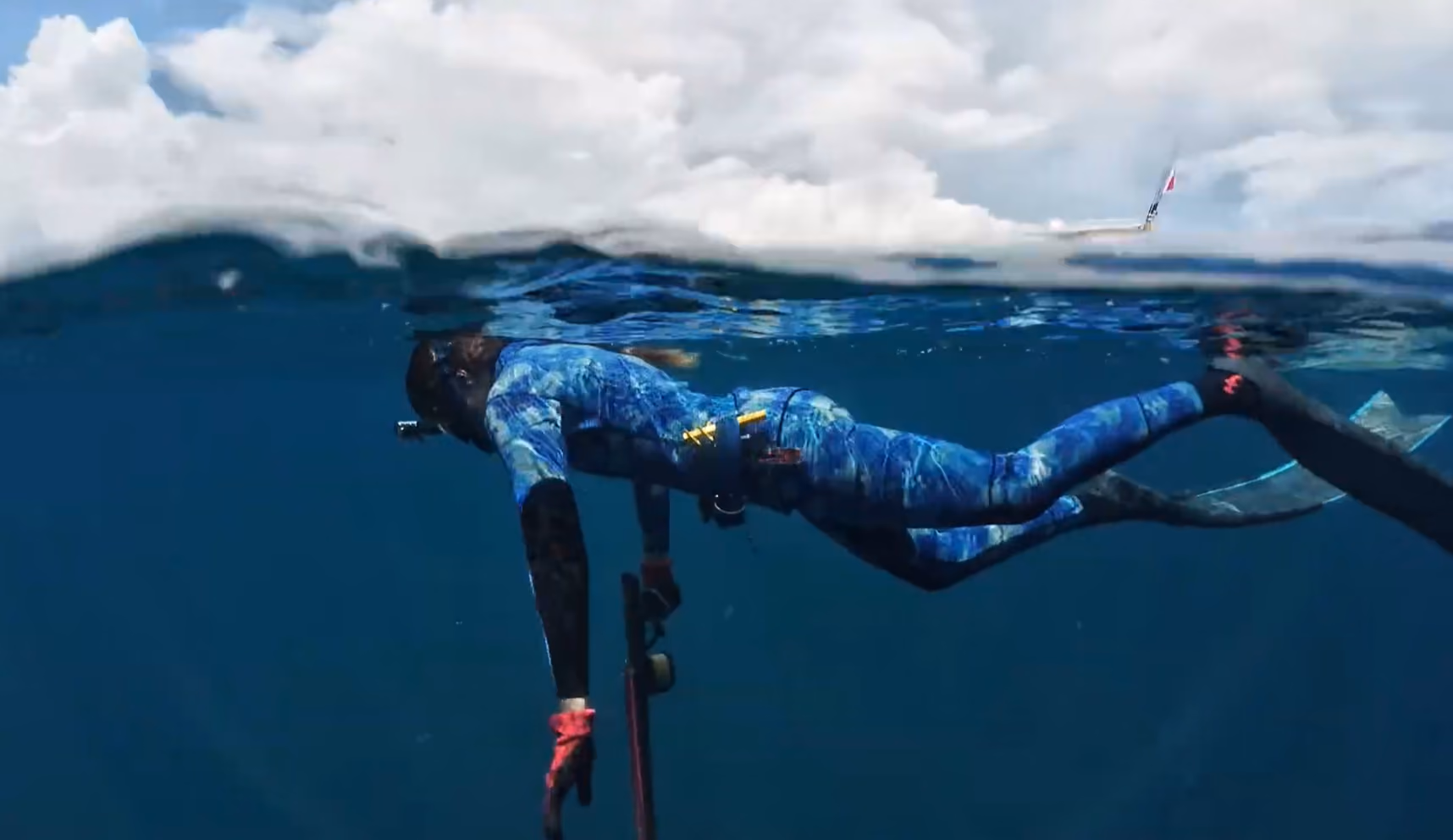 Person in blue camo wetsuit and fins swimming underwater with a speargun near the ocean surface.