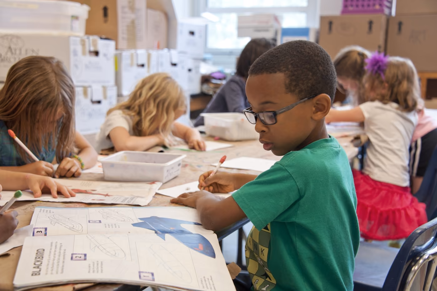 Young students seated at a table focused on drawing and writing activities in a classroom.