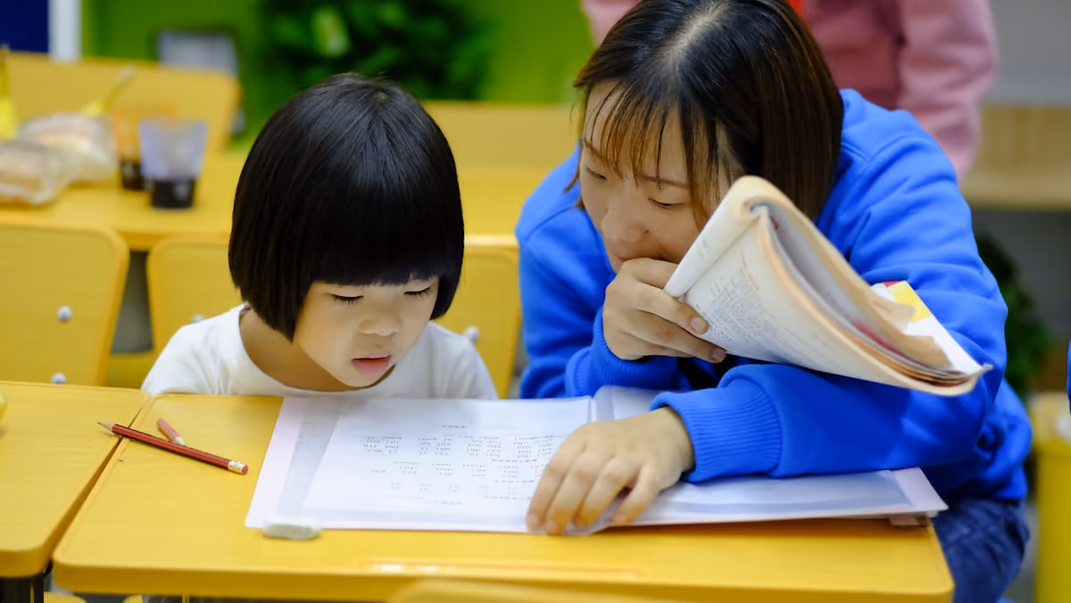 Teacher in blue jacket helping young girl with short black hair read a worksheet at a school desk.