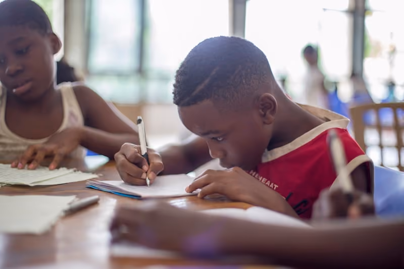 Young boy focused on writing in a notebook at a classroom table with other children.