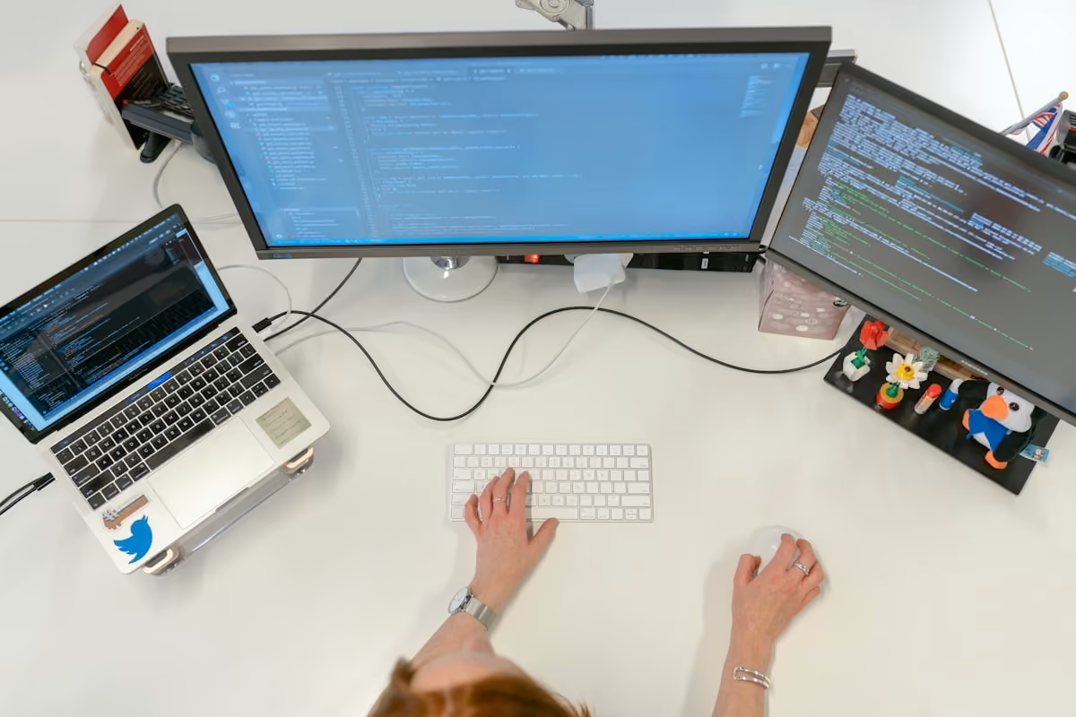 Person working at desk with laptop and two monitors displaying code and text editors.