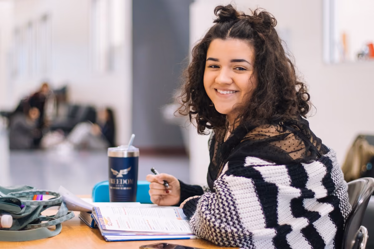 Smiling young woman studying with a pen, open book, and cup on table in a bright indoor space.