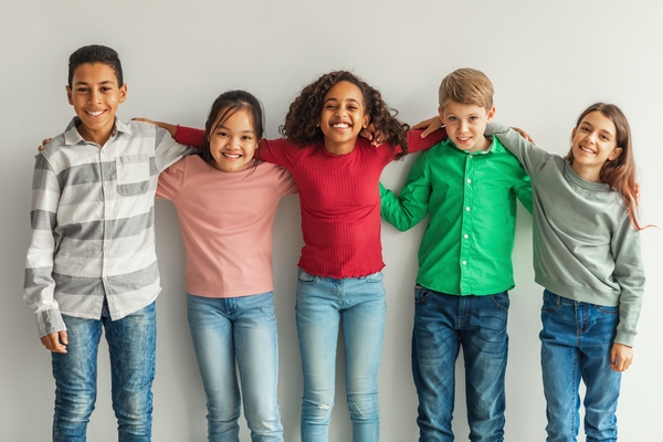 Five smiling children standing arm in arm against a plain wall, wearing casual clothes.