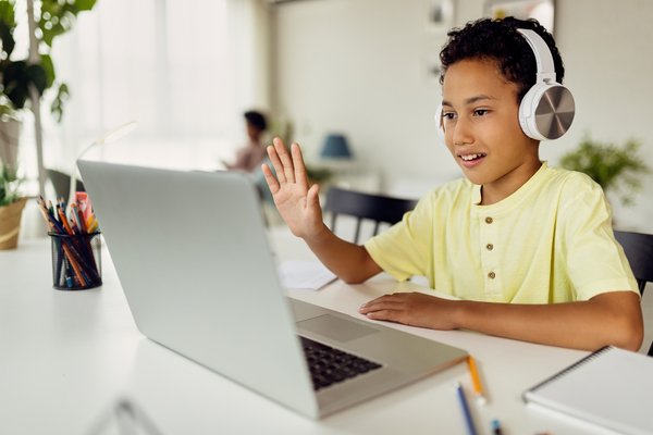Boy in yellow shirt and headphones waving at laptop during online video call at home.