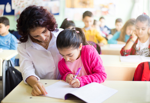 Teacher helping a young student write in a notebook in a classroom with other children.