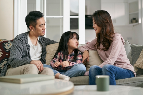 Asian parents sitting on a couch talking with their young daughter in a cozy living room.