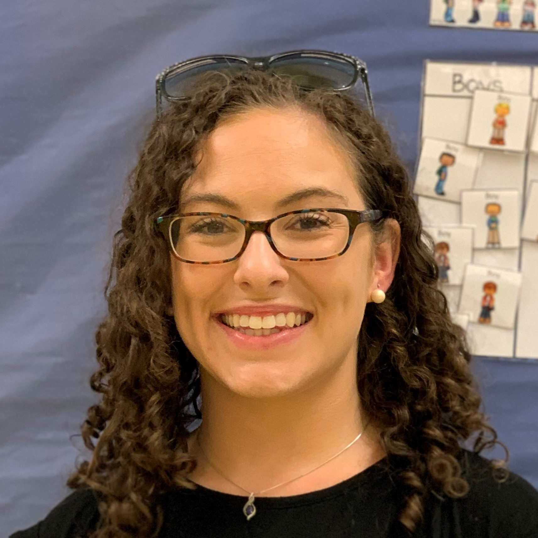 Smiling woman with curly hair, glasses, and pearl earrings in front of a blue bulletin board.