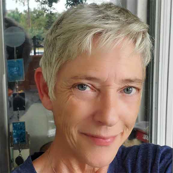 Close-up of a smiling person with short gray hair and blue eyes near a window.