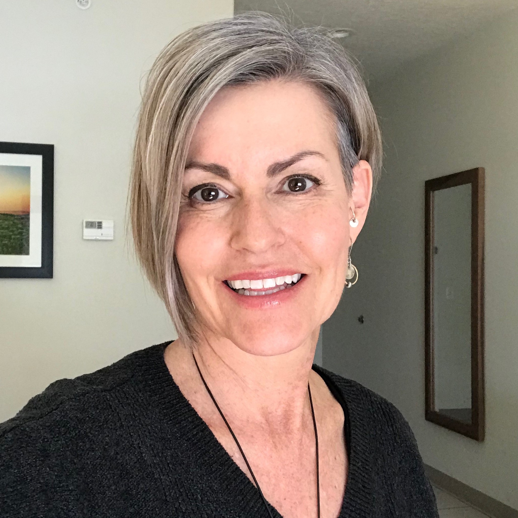 Smiling woman with short gray hair wearing black sweater and dangling earrings indoors.