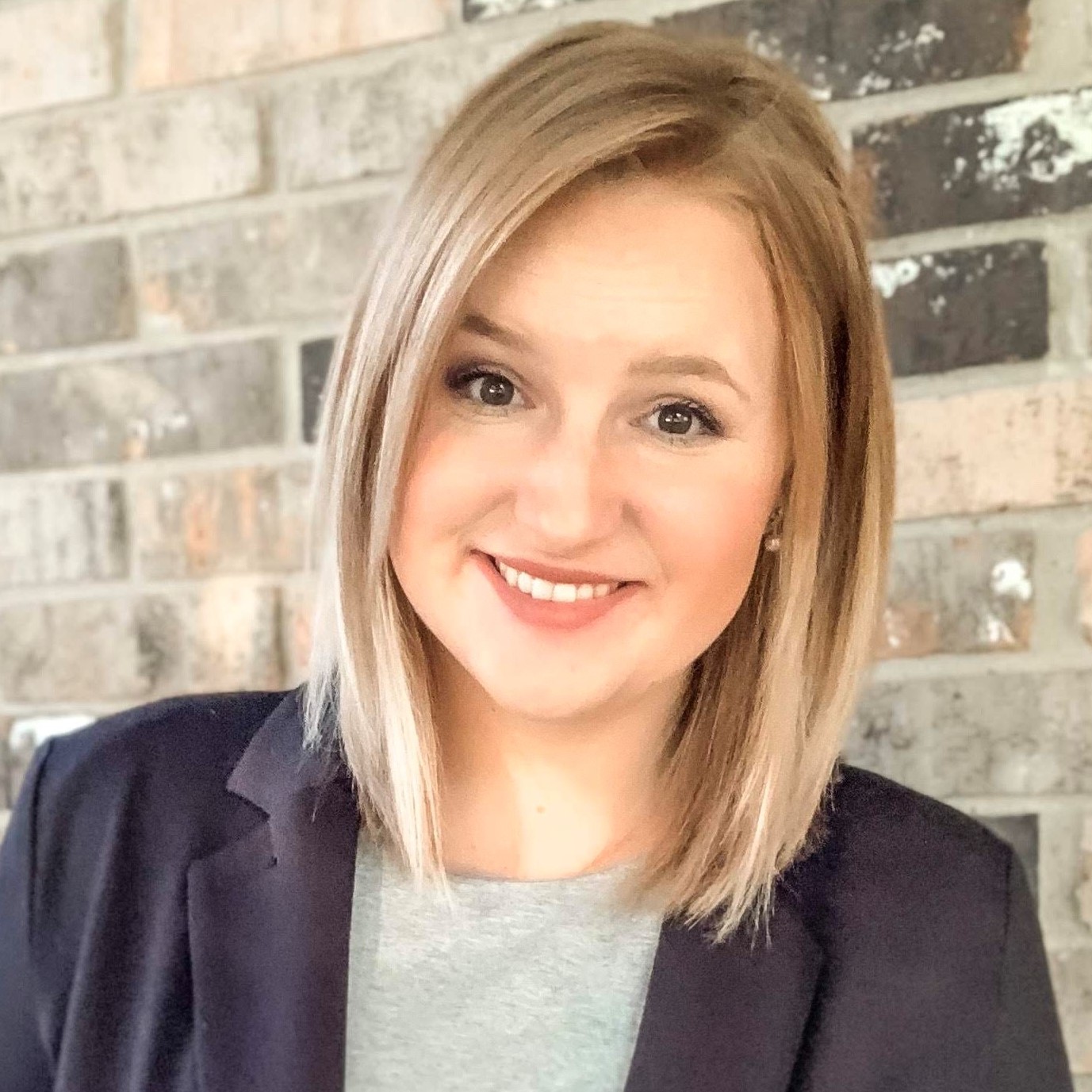 Smiling woman with blonde hair wearing a black blazer and gray top in front of a brick wall.