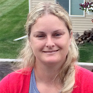 Smiling woman with blonde hair wearing a red cardigan and striped shirt outdoors near a house.