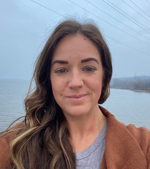 Woman with long brown hair wearing a brown coat and gray shirt stands outside near water under a cloudy sky.