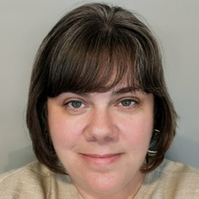 Woman with short brown hair and bangs, wearing a beige top, against a gray background.