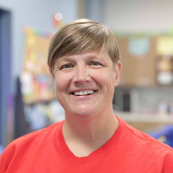 Smiling woman with short light brown hair wearing a red shirt against a blurred colorful indoor background.
