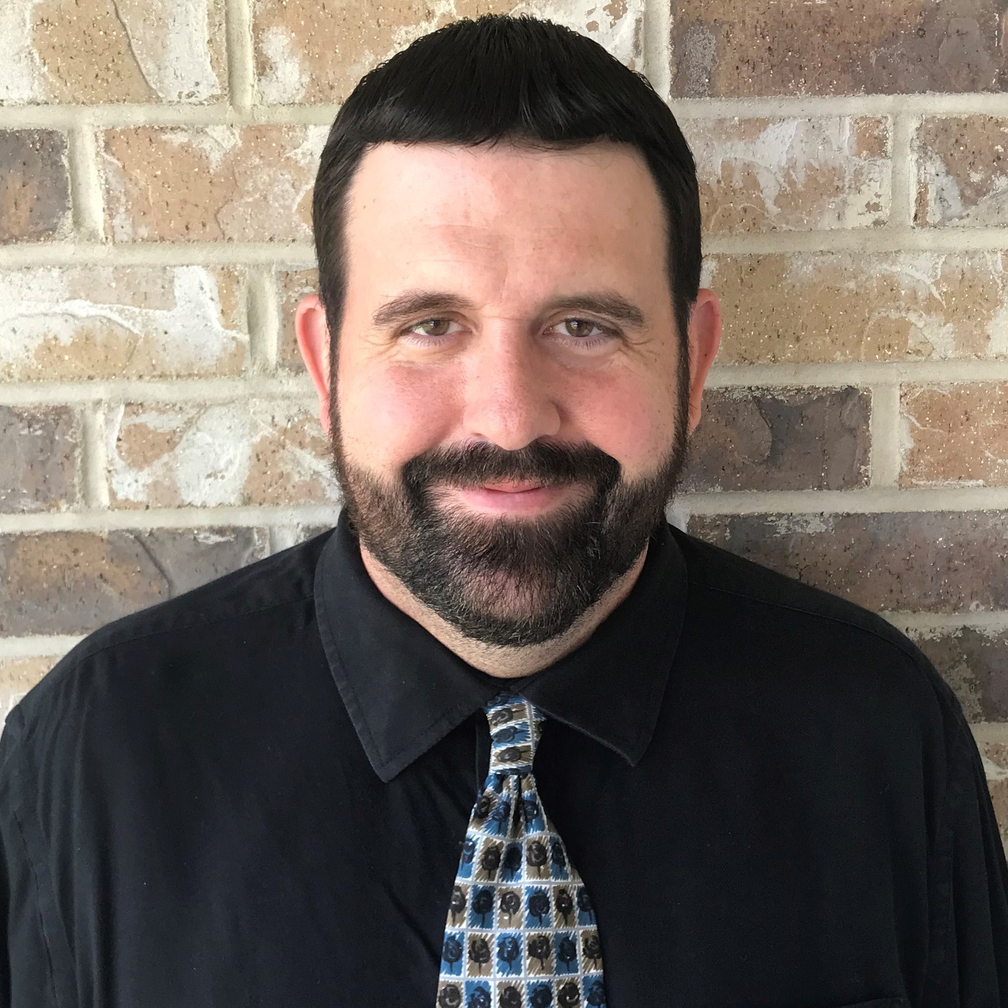 Smiling man with dark hair and beard wearing a black shirt and patterned tie against a brick wall.