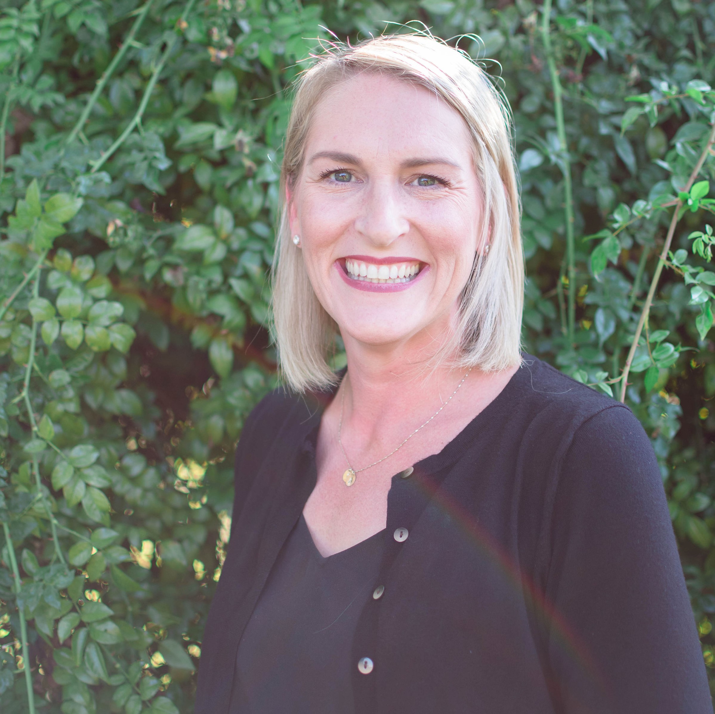 Smiling blonde woman wearing a black cardigan standing in front of green leafy bushes.