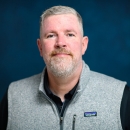 Man with short gray hair and beard wearing a gray fleece vest on a blue background.