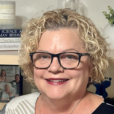 Smiling woman with short curly blonde hair and glasses in a home setting with books and photos behind her.