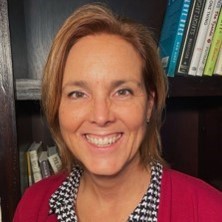 Smiling woman with short light brown hair wearing a red sweater and patterned collar in front of bookshelves.