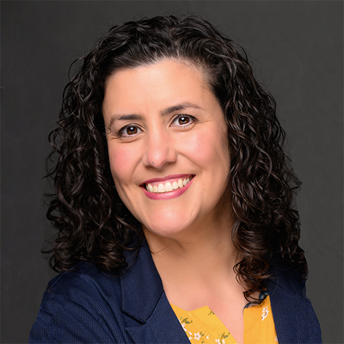 Smiling woman with curly dark hair wearing a navy blazer over a yellow floral top against a dark background.