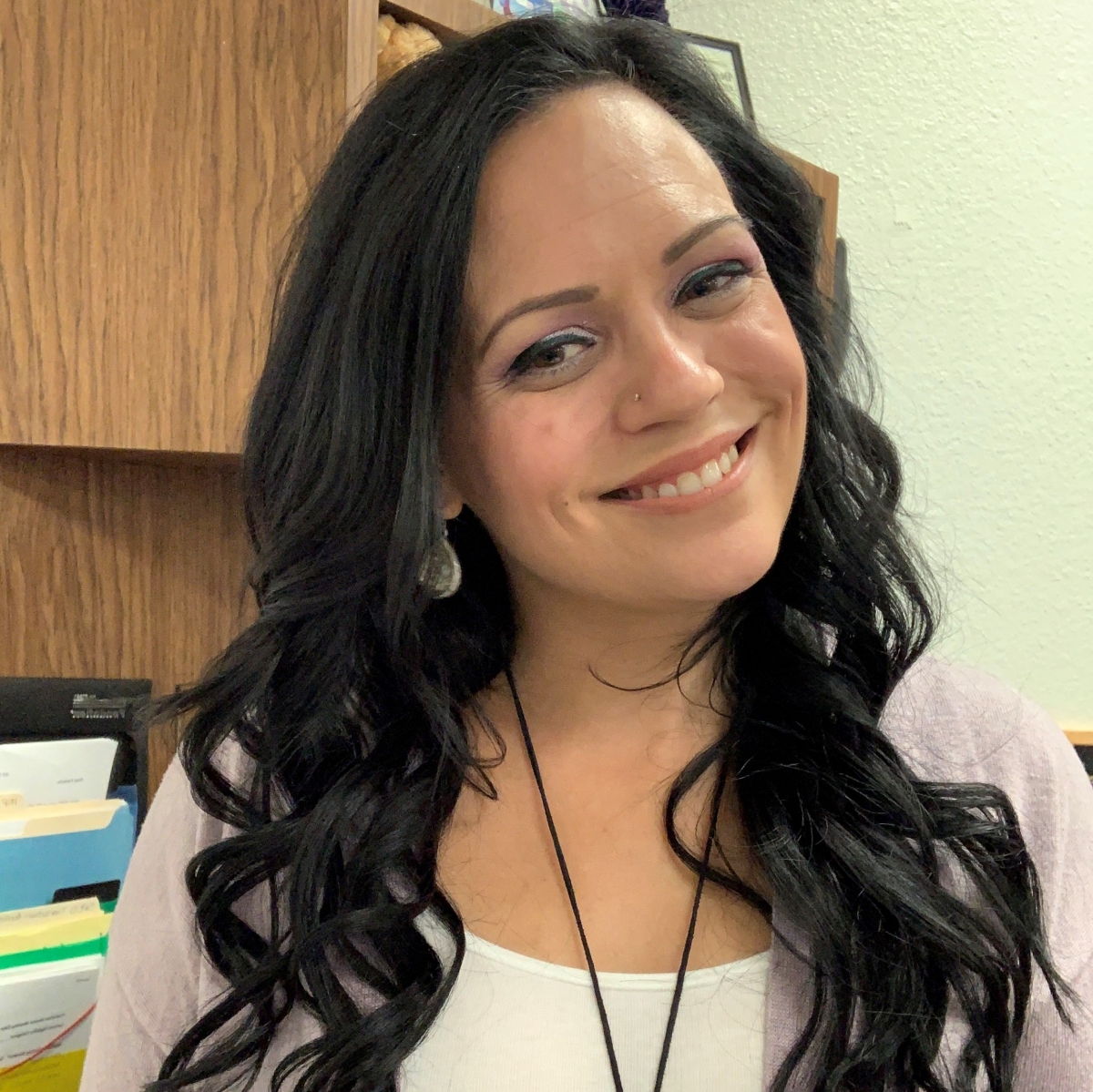 Woman with long black curly hair smiling, wearing earrings and a white top with a light cardigan.