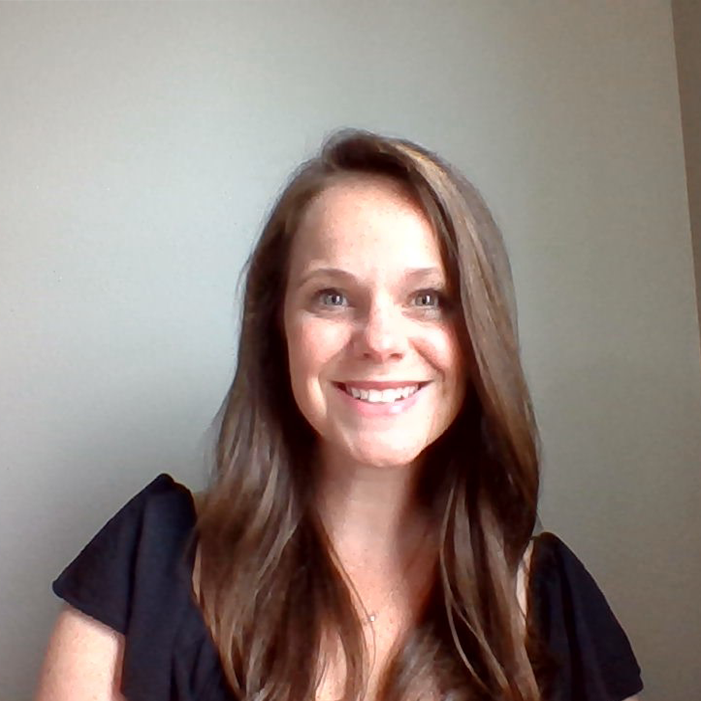 Smiling woman with long brown hair wearing a black top against a plain light gray background.