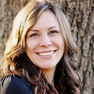 Smiling woman with long light brown hair standing outdoors against a tree bark background.