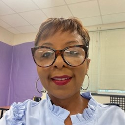Smiling woman wearing large glasses and hoop earrings with a purple background and window blinds.