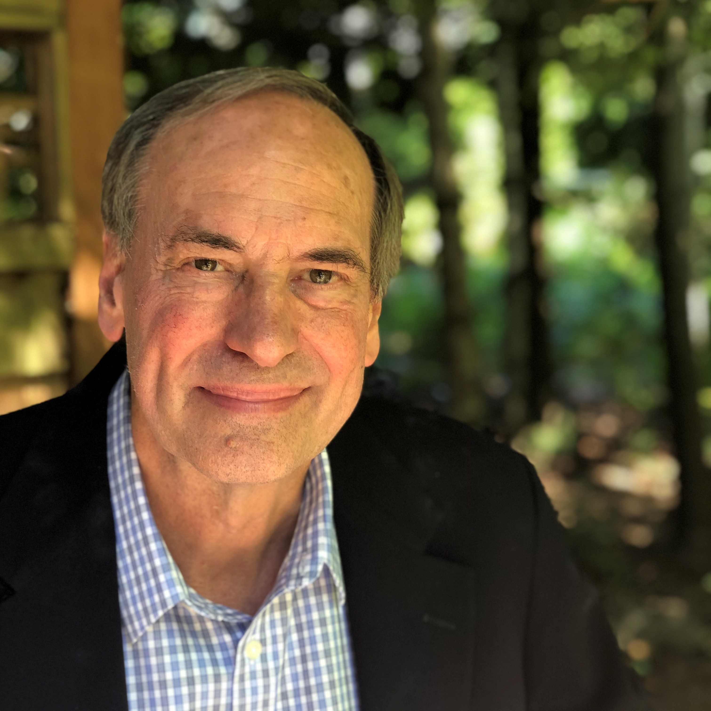 Smiling older man in a black blazer and blue checkered shirt outdoors with trees in the background.