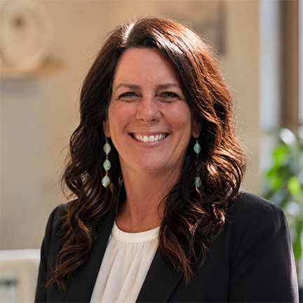 Smiling woman with long dark wavy hair wearing turquoise earrings and a black blazer over a white top.