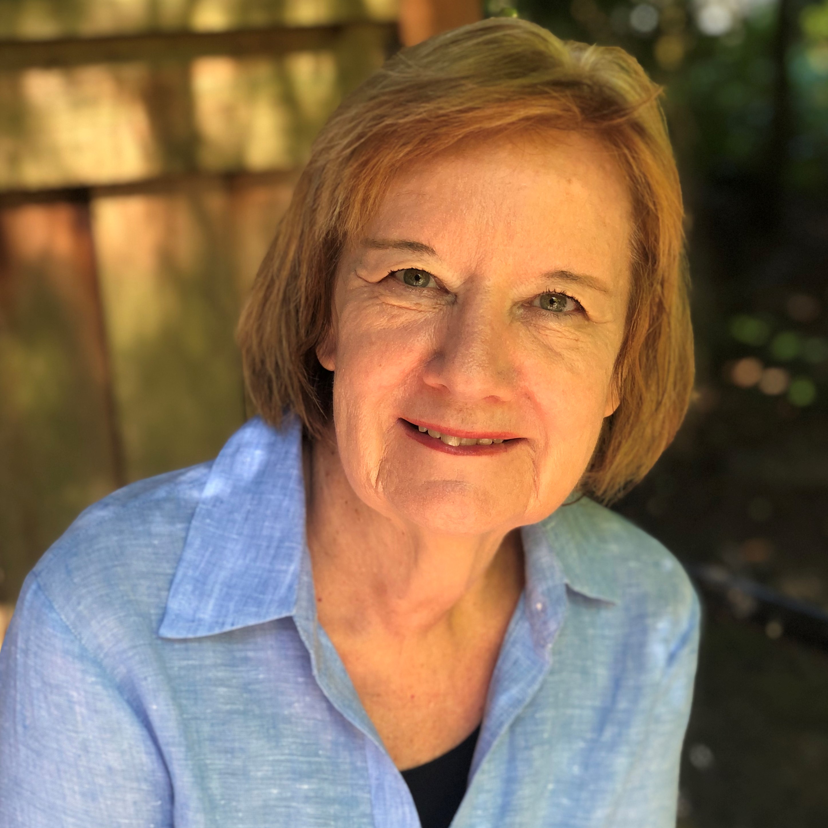 Smiling older woman with short light brown hair wearing a light blue shirt outdoors.
