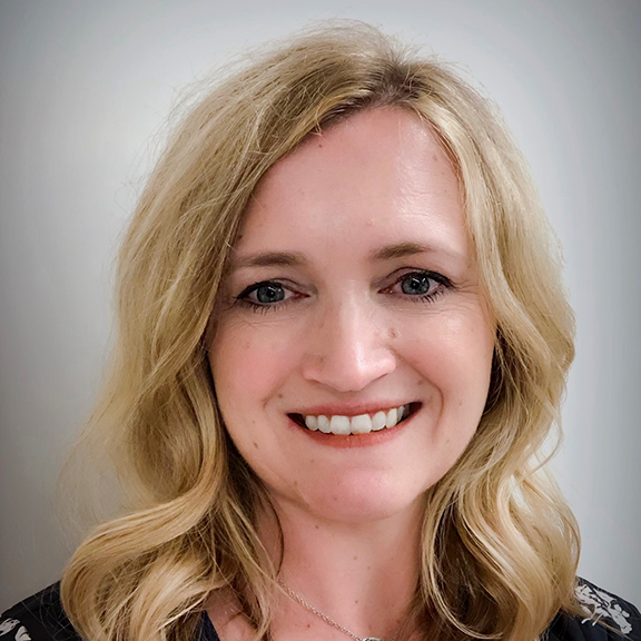 Smiling woman with wavy blonde hair wearing a black floral top and a silver necklace against a light background.