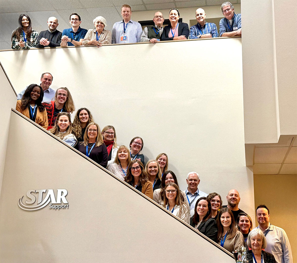 Group photo of diverse people standing on stairs and balcony inside a building with STAR Support logo.