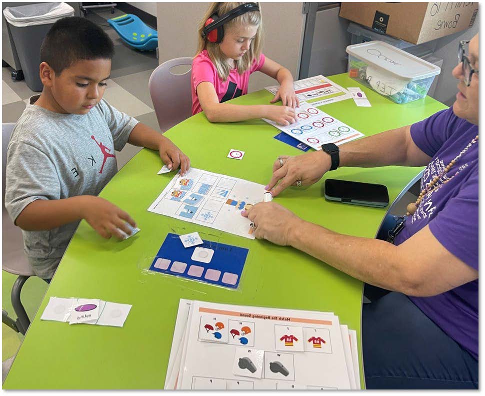 Two kids and an adult seated at a green table engaging in educational activities with cards and worksheets.
