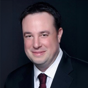 Portrait of a man in a dark suit with a white shirt and maroon tie against a dark background.