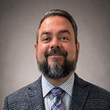 Smiling man with a beard wearing a plaid suit jacket, checked shirt, and striped tie against a gray background.
