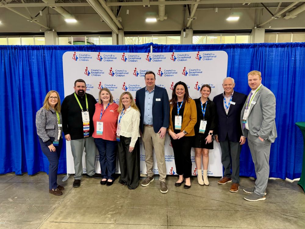 Nine people standing indoors in front of a Council for Exceptional Children backdrop with blue curtains.