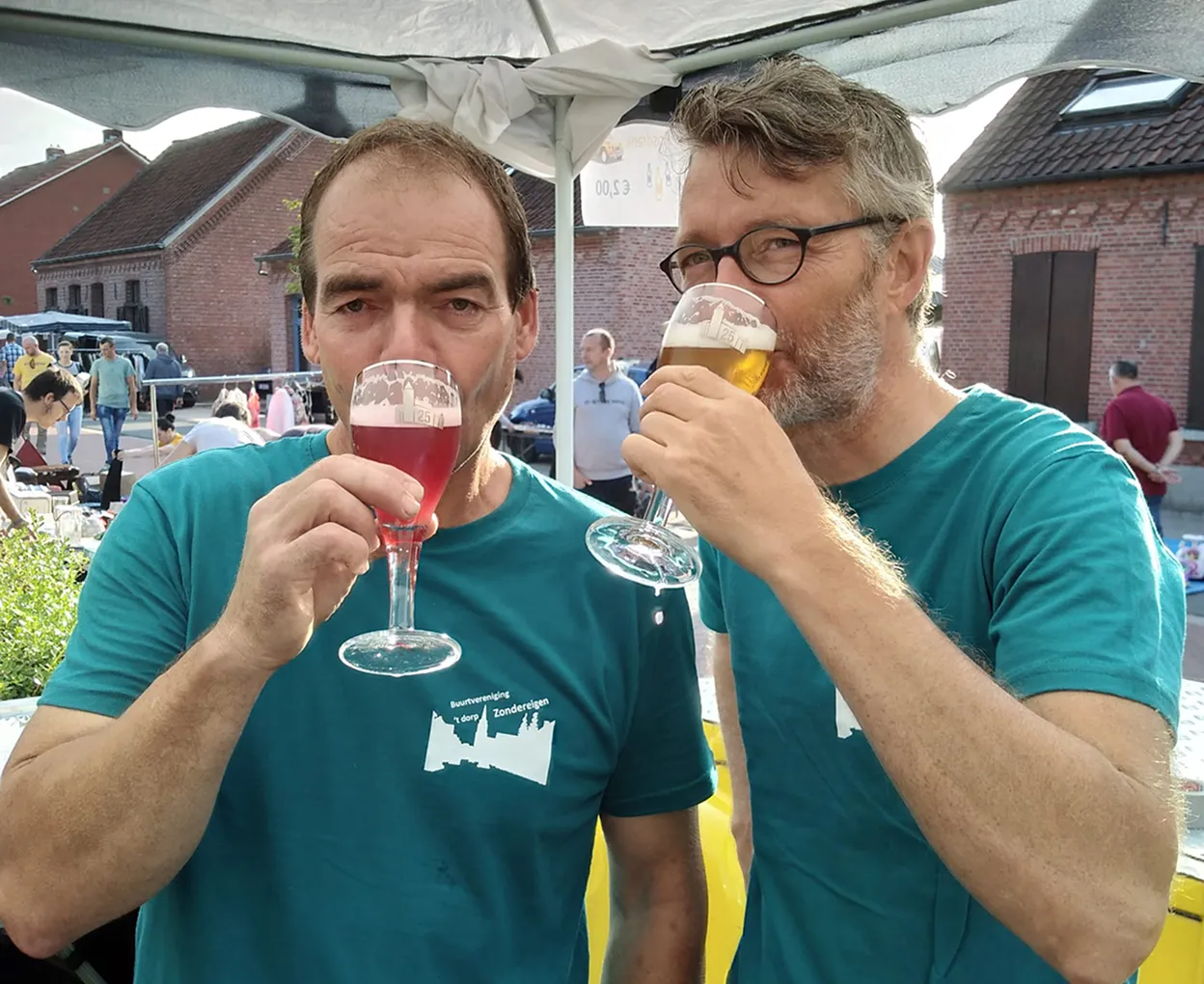 Twee mannen in groene T-shirts drinken rode en gouden bieren bij een buitenbijeenkomst.