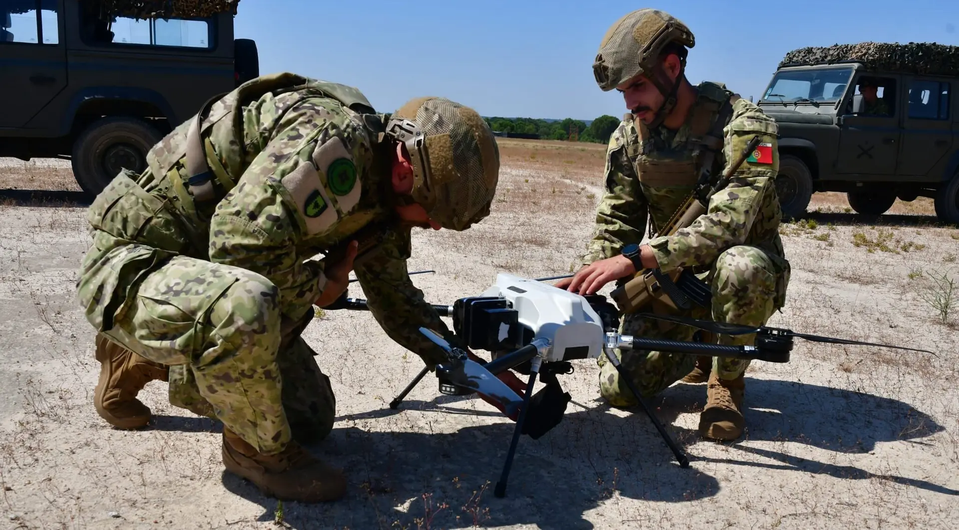 Two Portuguese soldiers in camouflage crouch over a BVQ418 quadcopter on dry ground, military vehicles behind them
