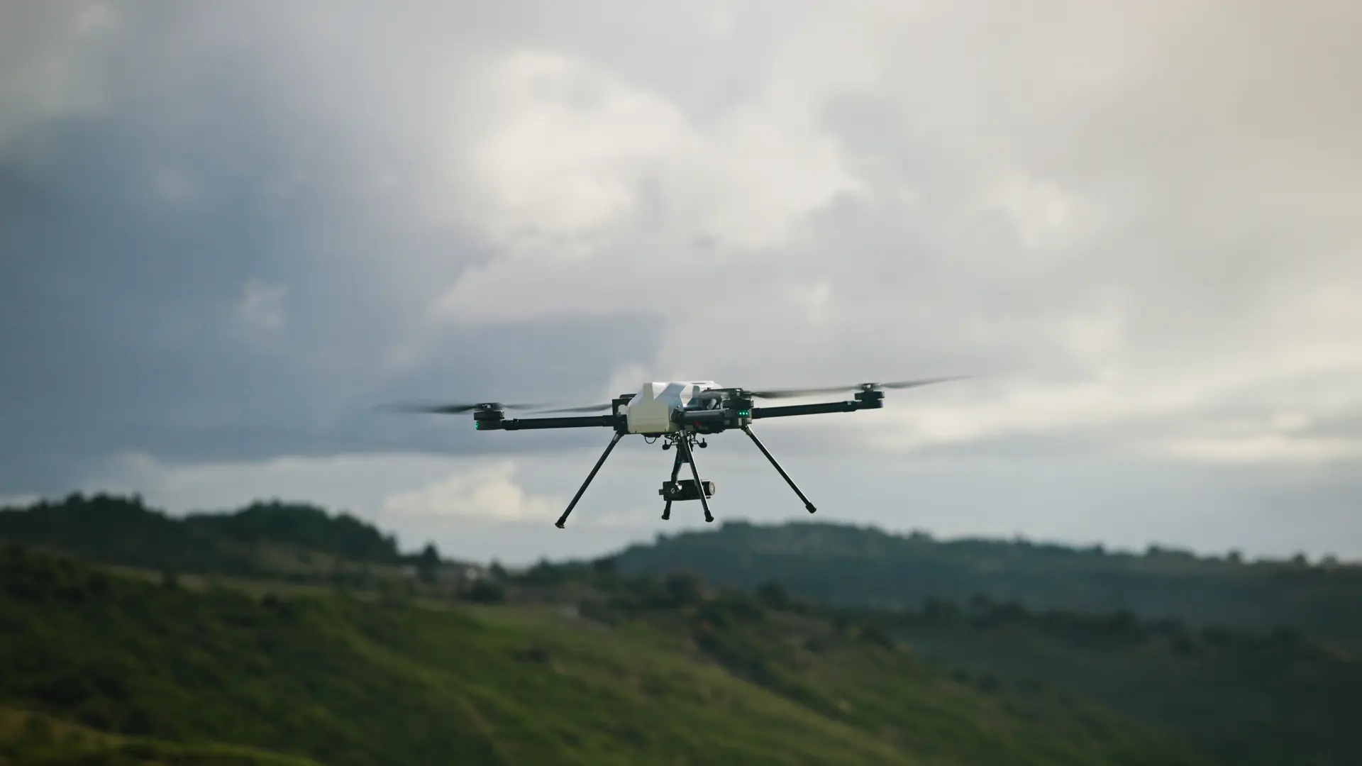 BVQ418 quadcopter hovering above green hills under a heavy overcast sky, gimbal camera mounted beneath the airframe