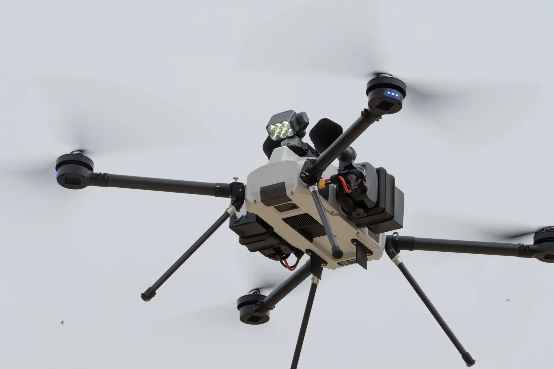 BVQ418 quadcopter in flight from below, LED spotlight and sensor payload on top, propellers blurred against a pale sky