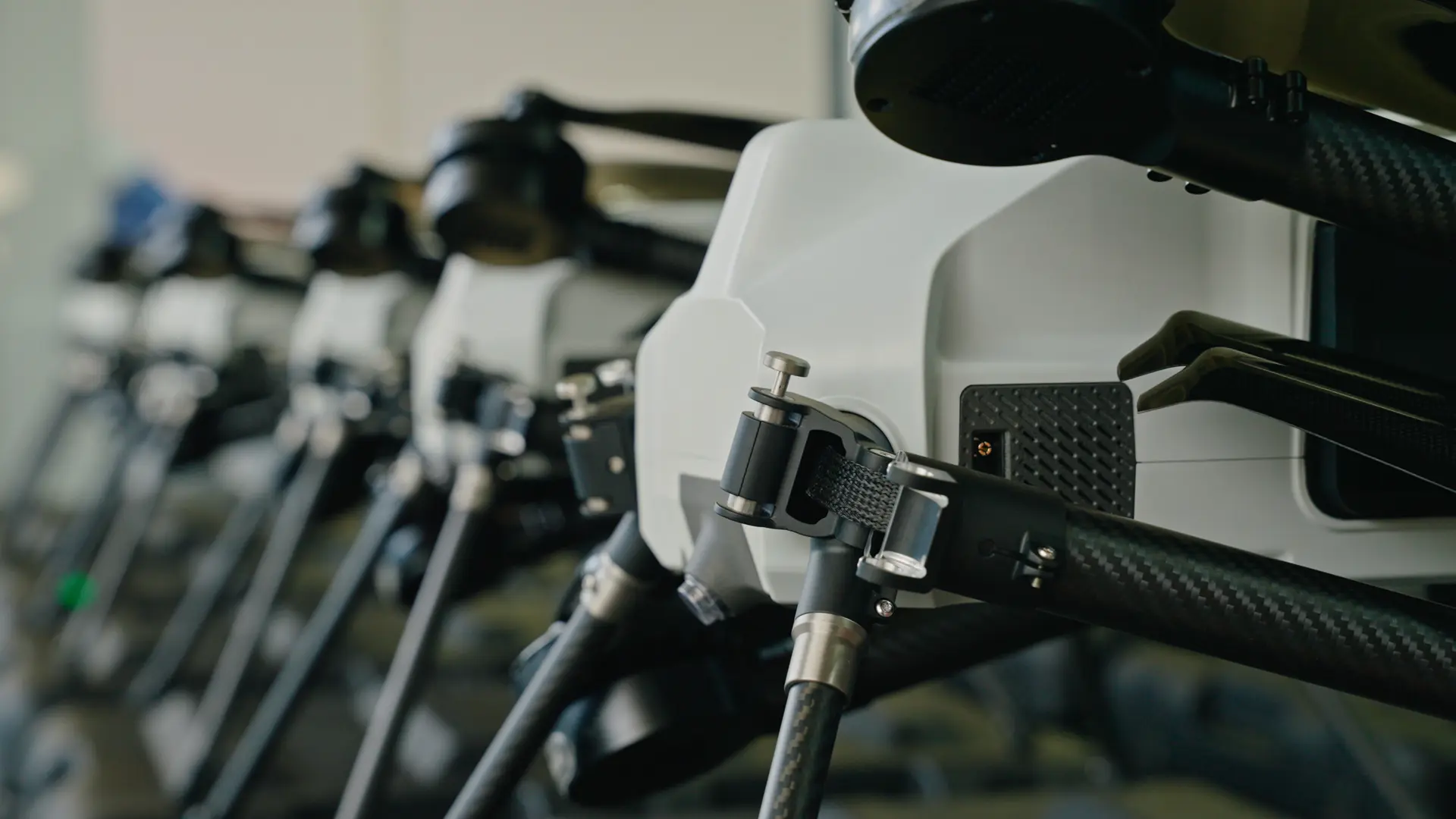 Row of BVQ418 quadcopters lined up indoors, close focus on a carbon-fibre arm with its clamp, strap and folding joint