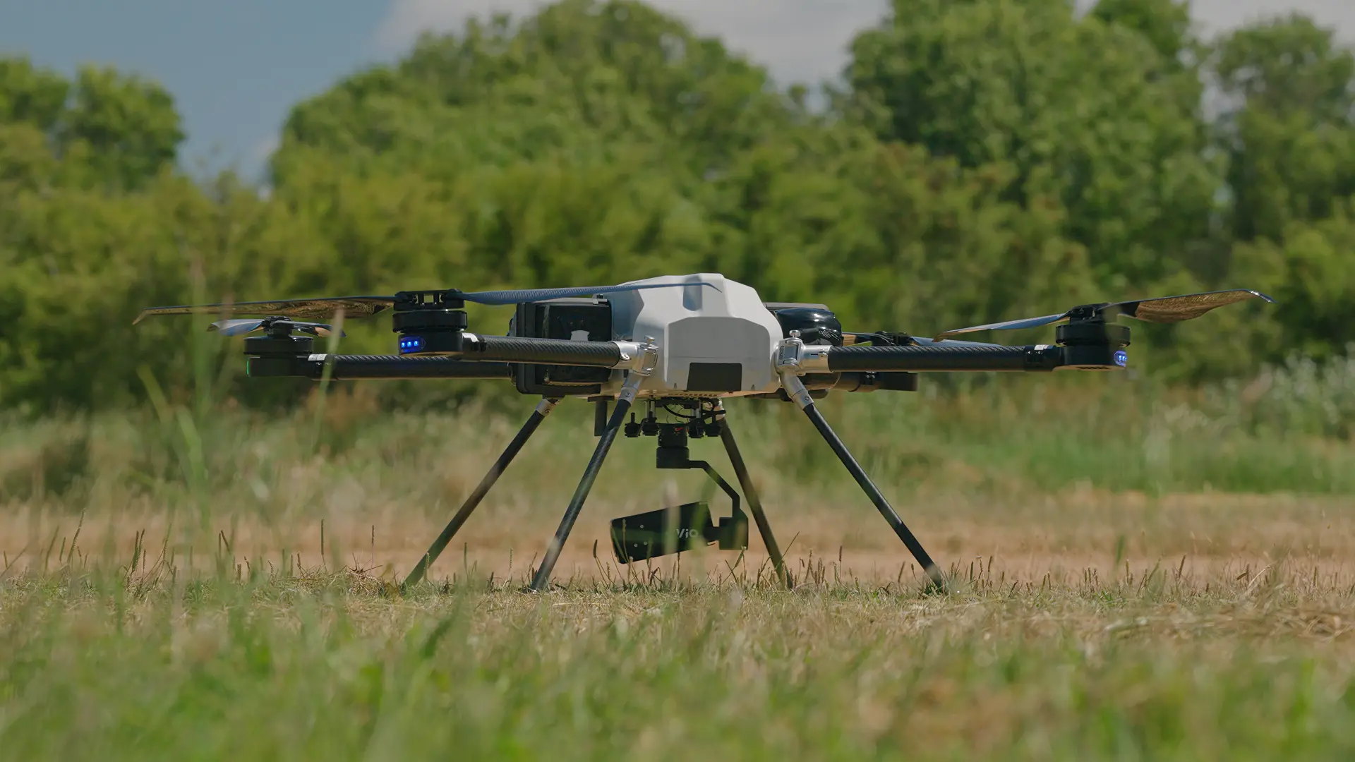 BVQ418 quadcopter standing on its landing legs in a grass field, gimbal camera mounted underneath, treeline in the background