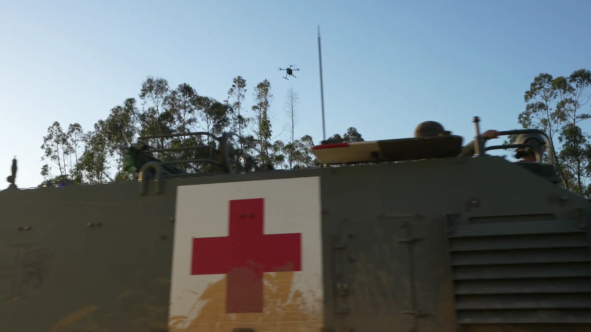 Armoured ambulance marked with a large red cross parked under eucalyptus trees, a BVQ825 flying high overhead