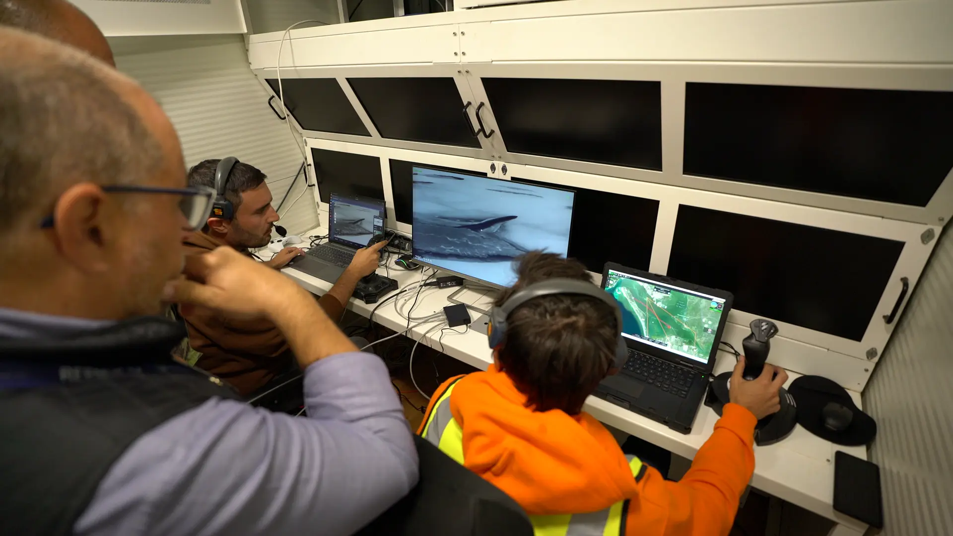 Three operators with headsets at laptops in a control cabin, screens showing aerial video over water and a mission map