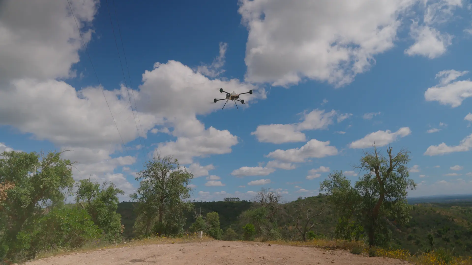 BVQ825 hovering above a wooded hillside under a bright cloudy sky, dirt track and power lines in the foreground