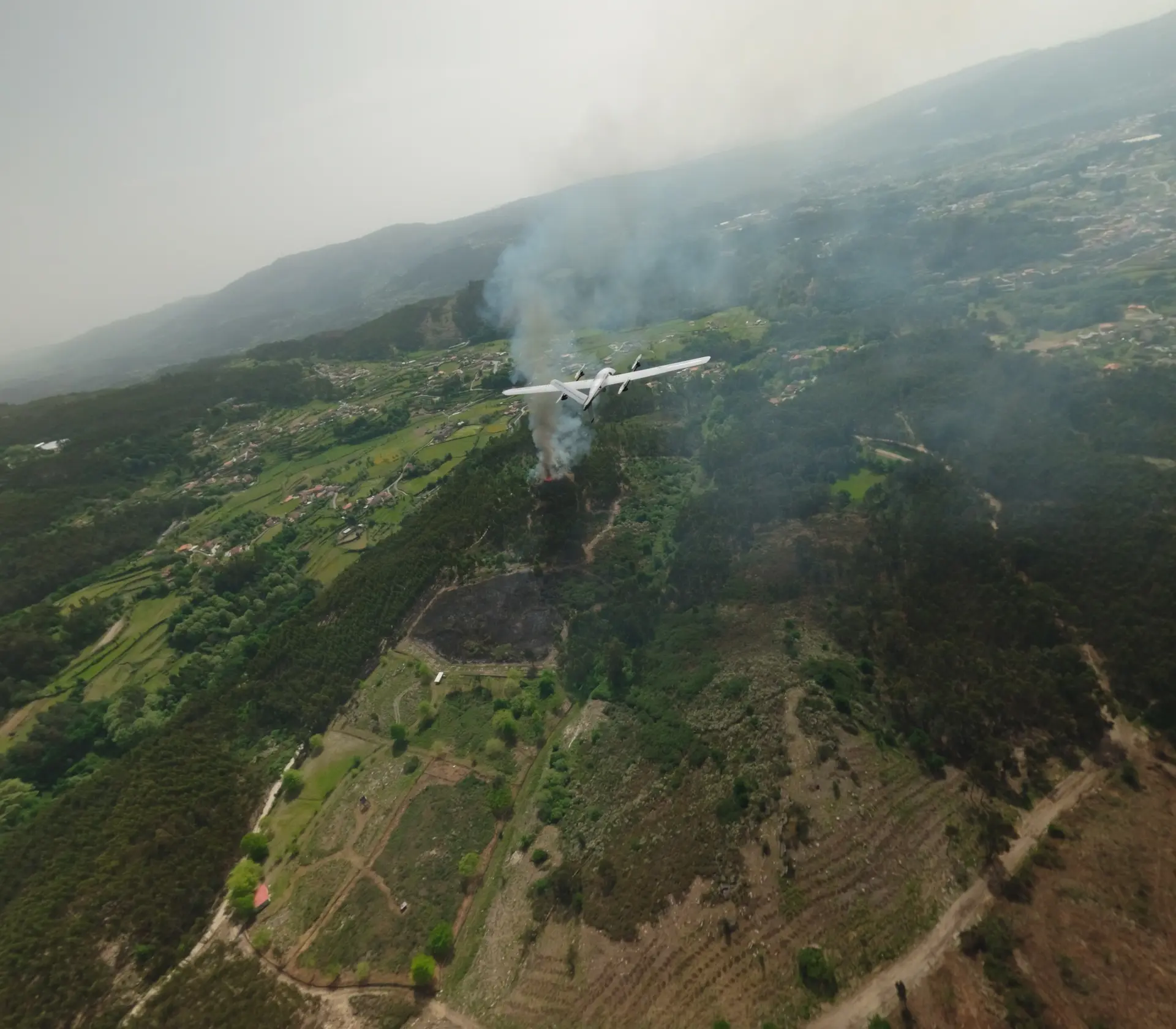 BVT516 fixed-wing drone banking above a wooded valley where smoke rises from a fire, villages and burnt ground below