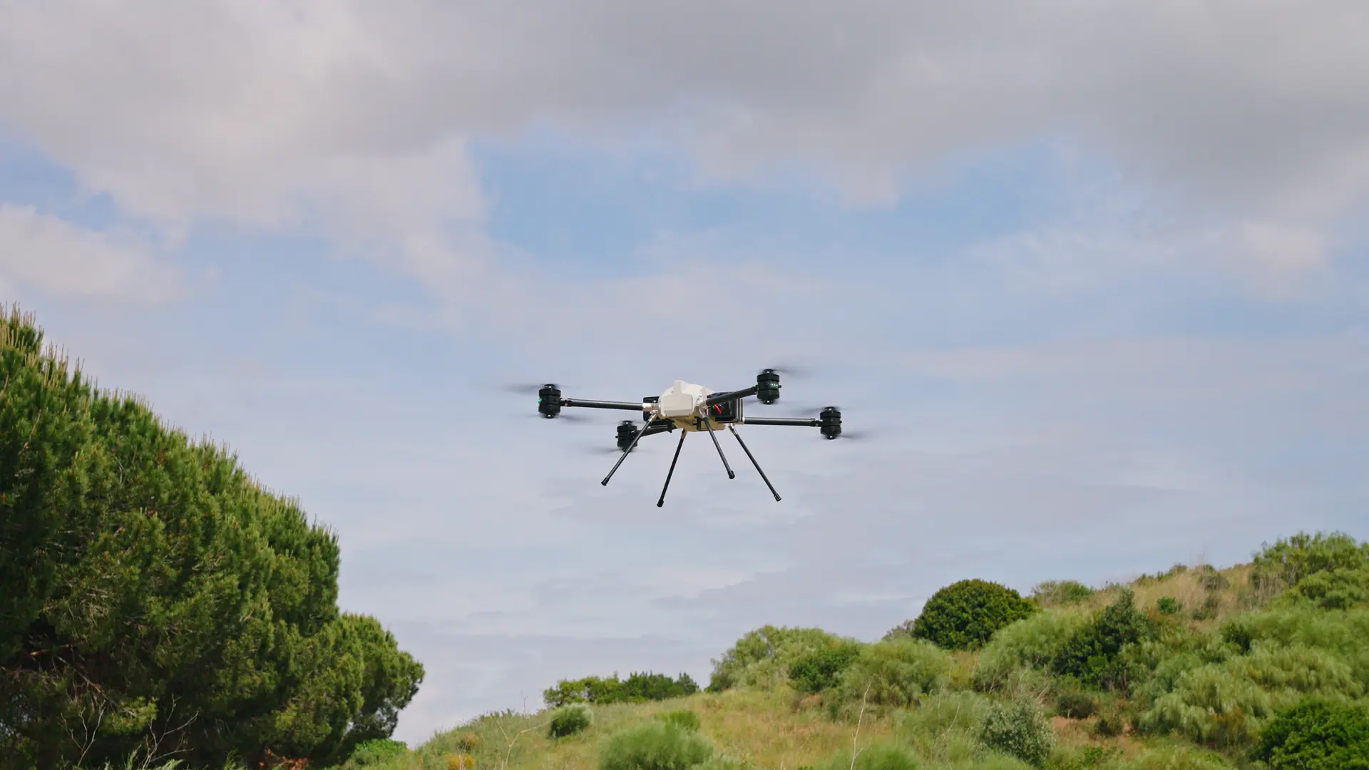 BVQ825 hovering above coastal scrub and pine trees under a pale cloudy sky, landing legs extended